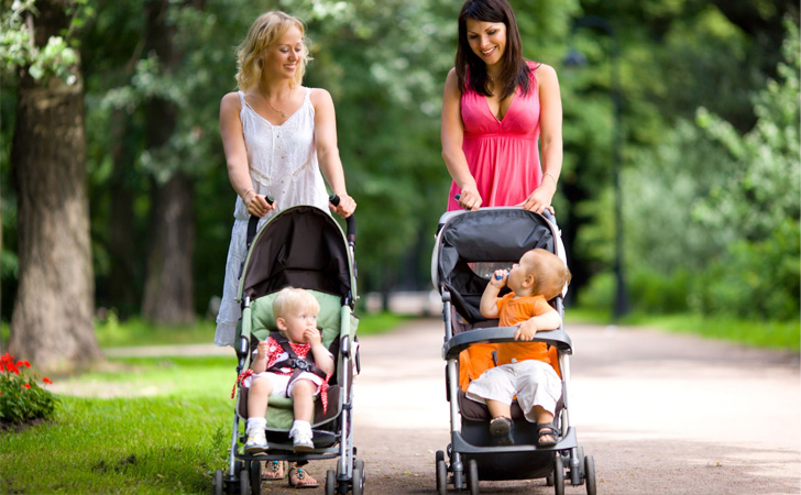 Dwie mamy spacerujące w parku prowadzące spacerówki, w których znajdują się ciekawe świata maluchy